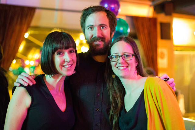 Three smiling individuals posing together at a festive event, with colorful balloons in the background, representing community engagement at the Love Bomb 2017 fundraiser for My Voice Music.